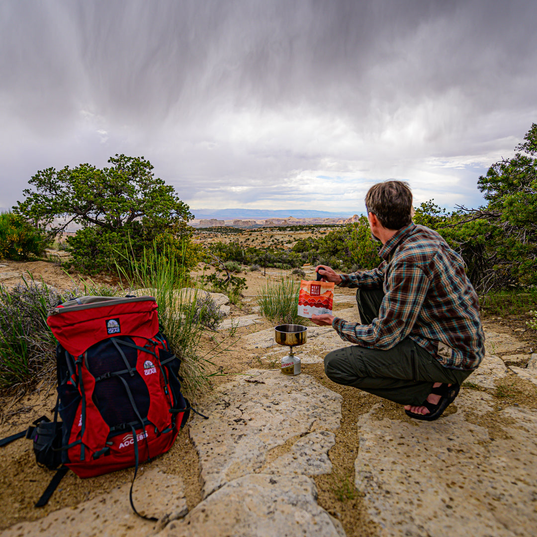 a backpacker makes a meal on a rock while looking out at a vista