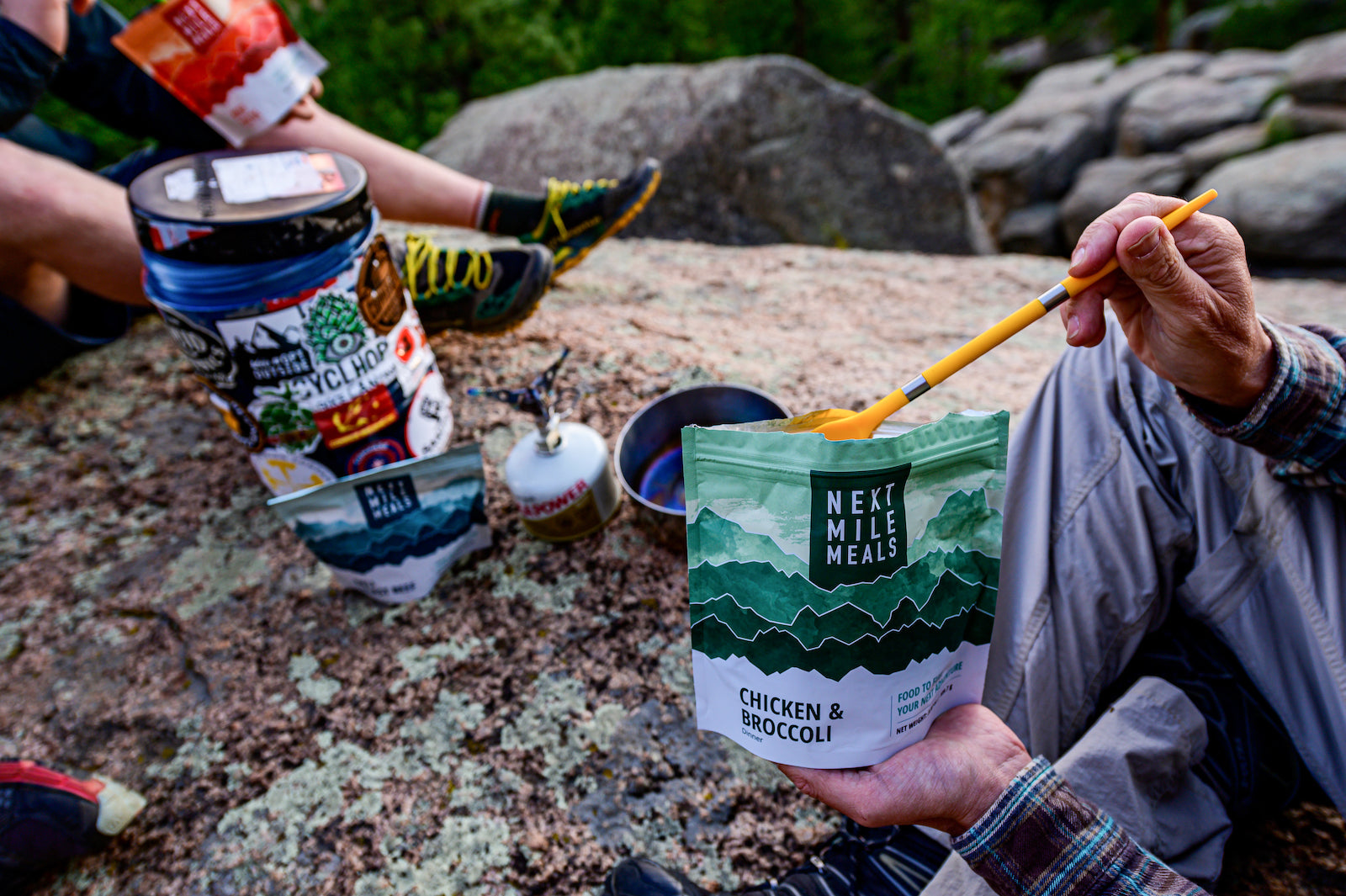 A backpacker holding a Next Mile Meals Chicken & Broccoli bag while sitting on a rock