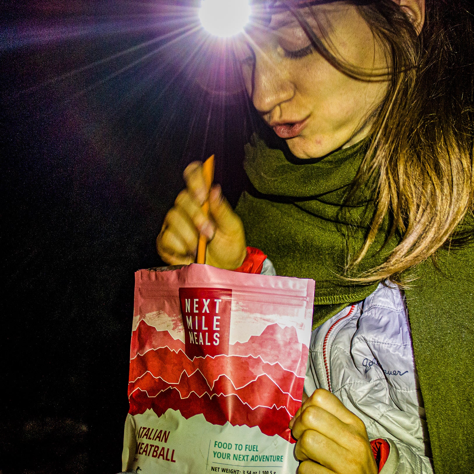 A woman eats a backpacking meal in the dark lit by a headlamp