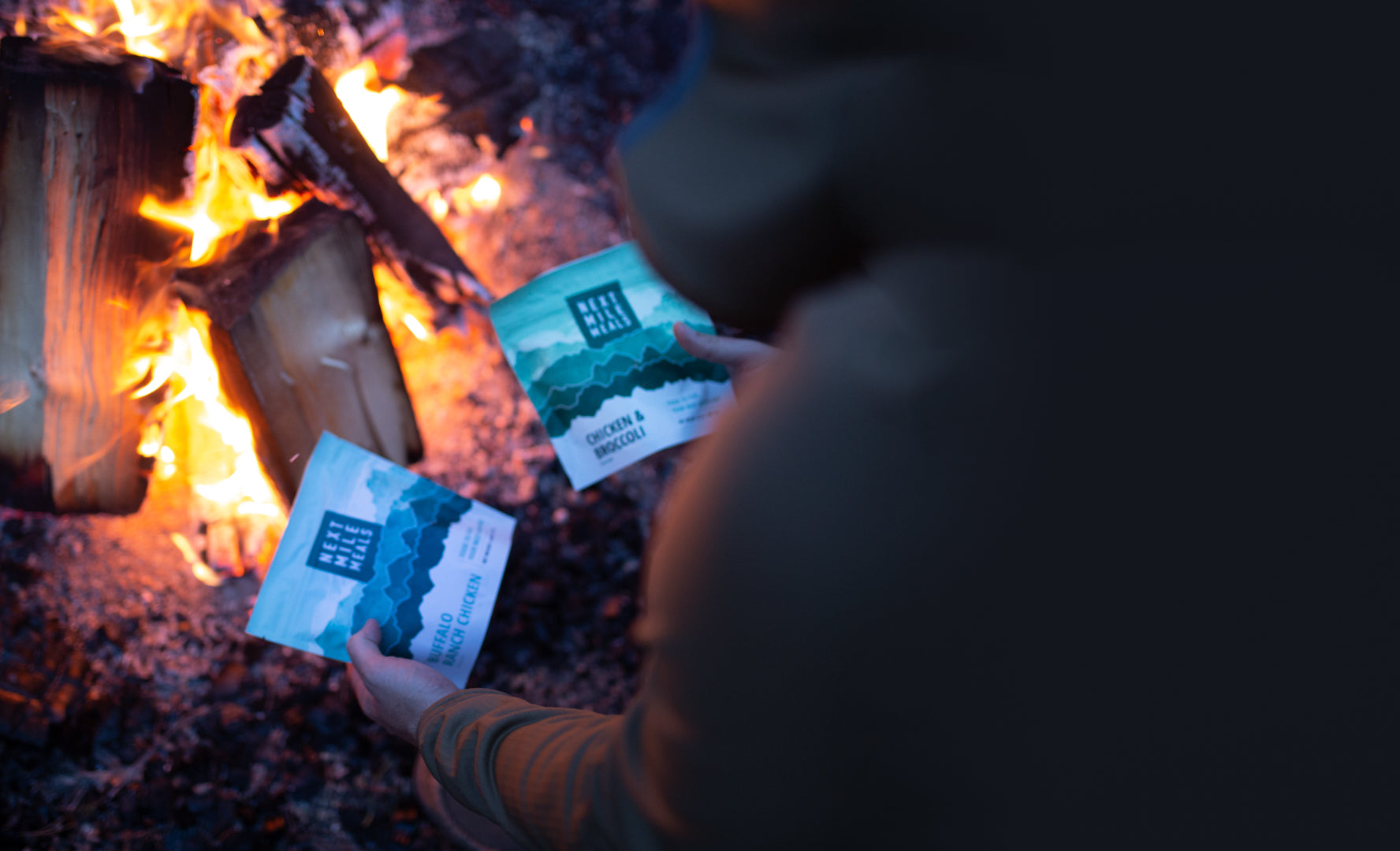 a person holding two backpacking meals in front of a fire.